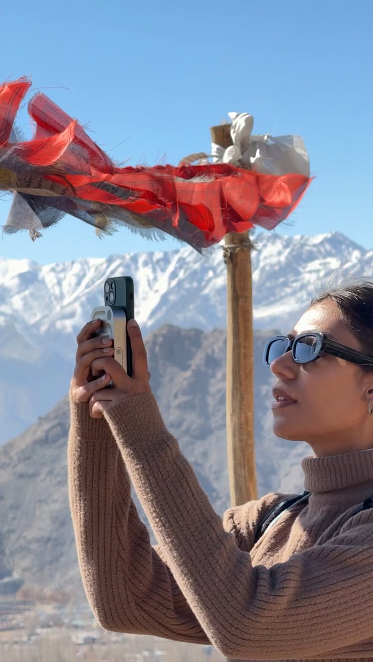 A woman wearing sunglasses and a brown sweater holds a phone up, positioned in front of her face. The background shows a clear blue sky, snow-capped mountains, and colorful prayer flags on a wooden pole. The composition focuses on the woman's profile, with the phone and a mountain setting providing context.