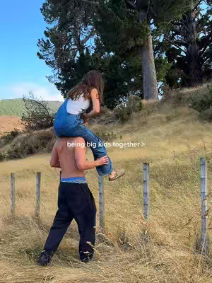 A couple is outside embracing in a happy moment. The man is carrying the woman on his shoulders while walking through a grassy field near a fence. The image has a natural color palette with green, yellow, and blue tones.