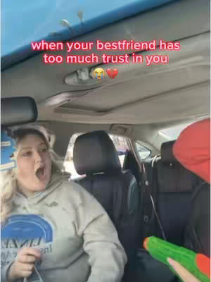 The cover image captures a scene inside a car. A woman with blonde hair, wearing a grey hoodie, is in the front seat with her mouth wide open in shock. Another person, partially visible, is in the back seat. A green water gun is pointed towards the woman. The background shows the car's interior with the roof and seats.