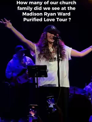 Shot of a woman on stage with arms raised, singing. She is wearing a cap and a white t-shirt. The background is dark, possibly a concert venue.