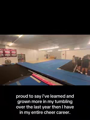 A view inside a gym where cheerleaders train. The camera is at a low angle, showing a large, blue mat in the center, and further back there is a trampoline. Several people, likely cheerleaders and coaches, are standing around. The lighting is bright, coming from overhead fixtures.