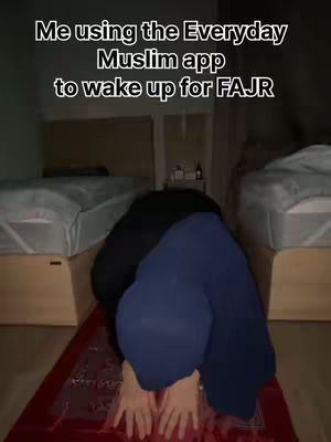 A person, presumably a Muslim woman, is shown on a patterned prayer mat kneeling on the floor. The person is praying, their back to the camera. There are two beds in the background with white sheets and pillows on either side of the person. The person is wearing a dark outfit.