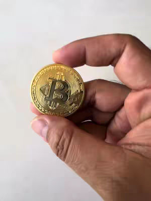 A person holding a gold Bitcoin coin, with the coin prominently displayed in the center of the image. The person's hand is partially visible, holding the coin against a plain white background.