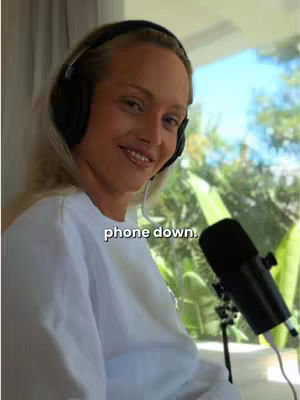 A woman with blonde hair wearing a white sweatshirt and headphones, smiling towards the camera. A microphone is to her right, and a window is visible in the background.