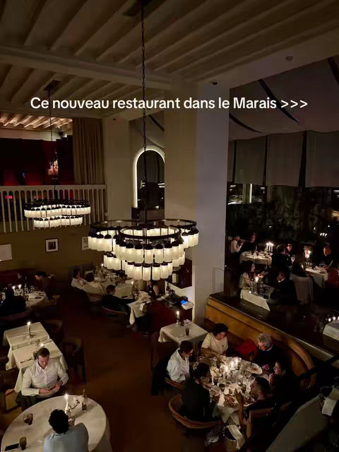 An overhead view of a dimly lit, upscale restaurant interior. The room is dominated by dark wood and warm lighting, with multiple tables occupied by patrons. Large, ornate chandeliers hang from the high ceilings. Across the background, a balcony overlooks the dining level. The overall aesthetic evokes a sense of elegance and exclusivity.