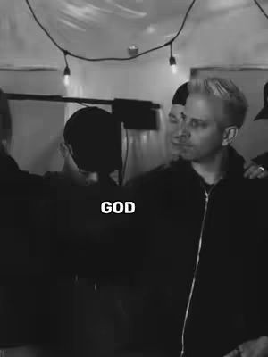 A black and white image featuring a group of men. The man in focus is fair-skinned with light hair and a black jacket, positioned on the right side of the frame. Beside and behind him are other men, blurred, and a portion of a lighting rig is visible on the left. A string of lights hangs from the ceiling. The word 'GOD' is overlaid in bold white letters.