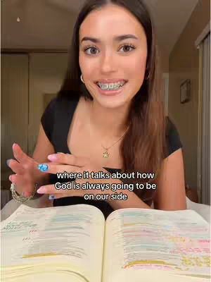 A young woman with long brown hair, wearing a black top and braces, is sitting in front of an open book. She is smiling and gesturing with her hands. The background includes a room with a dark-colored door.
