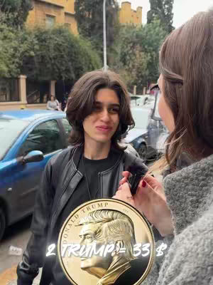 A woman holds a large gold coin featuring a profile of a man, likely Donald Trump, while talking to another person. The background includes a street with trees and parked cars.