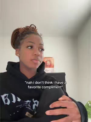 A young woman with braided hair is captured in a selfie-style video. She's wearing a black hoodie, and her expression is thoughtful. Text overlaid on the image reads, "nah i don't think i have a favorite compliment". The background includes a light-colored wall and a glimpse of an object, possibly a painting or a decorative item, adding some context to the setting.