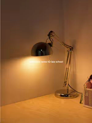The image is a close-up of a desk illuminated by a desk lamp. The lamp, with its metallic structure and a black dome, casts a warm glow on the surface. The composition shows a study setting with a corner of a wooden desk and a wall. On the desk, there are items like a pen holder in the right corner, suggesting a study or work environment.