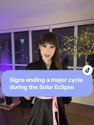 A woman with long dark hair and bangs is centered in the frame, wearing a black top and a pink ribbon around her waist. A light blue oval with white text indicates the video topic. The set has large windows overlooking a night cityscape.