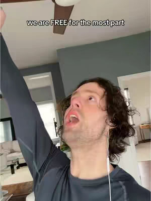 A man with brown curly hair is looking up with his mouth open and one arm raised up in the air. The background features a ceiling fan and neutral colored walls.