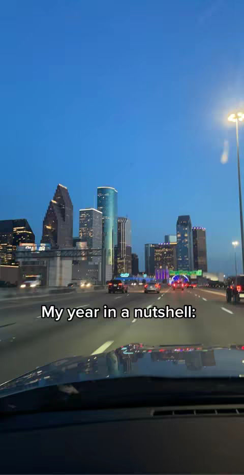 A scenic view from a car at dusk, likely a drive through a city. The sky is deep blue, with city lights beginning to illuminate buildings and roadways. The foreground includes the car's dashboard, adding a sense of being in motion. Text overlayed at the bottom says, "My year in a nutshell:".