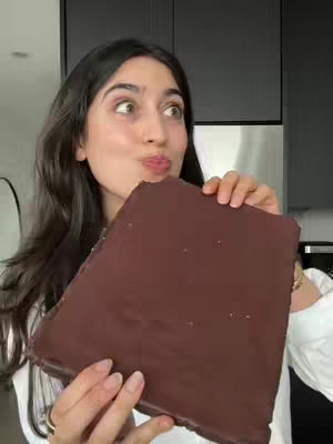 A woman with dark hair holds a large, rectangular piece of dark chocolate up to her face, looking slightly surprised with her lips pursed. The background includes a modern kitchen with dark cabinets.