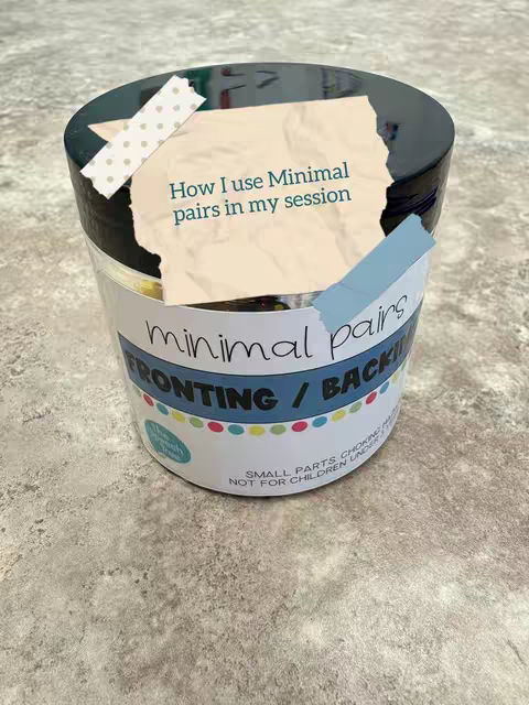 The image showcases a jar of minimal pair mini objects, with a custom label and a handwritten note taped to the lid. The jar sits on a textured gray surface, likely a tabletop or countertop.