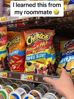 The image shows a grocery store shelf filled with bags of chips. In the foreground, there are bags of Cheetos in bright orange and red packaging. A hand is visible on the right side of the frame, presumably reaching for or pointing at the chips. The background is filled with other snack products.