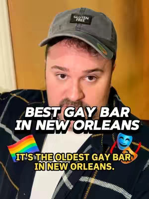 A man wearing a baseball cap and a plaid shirt is in a selfie-style shot. The image has overlaid text that says "BEST GAY BAR IN NEW ORLEANS"; below this is another text element that reads,  "IT'S THE OLDEST GAY BAR IN NEW ORLEANS." There's a small rainbow flag, a theater mask, and a smiling mask graphic.