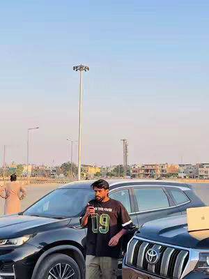 A man is standing next to a car, appearing to use his phone. There's a cityscape and sky in the background.