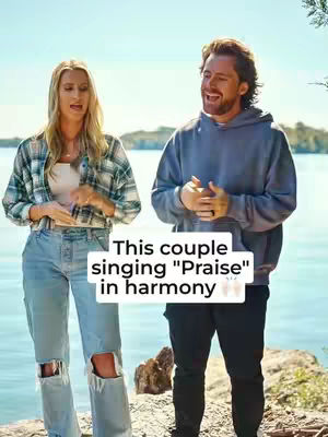 A couple stands facing the camera with a natural background. The woman on the left has blonde hair and is wearing a plaid shirt and jeans. The man on the right has short brown hair and is wearing a gray hoodie and black pants. Overlaying them is text stating 'This couple singing "Praise" in harmony'.