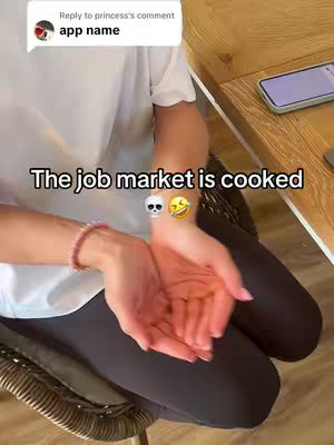 The image shows a person from the waist down, seated and holding open hands in front of them. The background is a wooden surface and a cellphone. Text overlays read, with a speech-bubble indicating that the app name is 'app name' and the main context says 'The job market is cooked' with skull and laughing face emojis. The person is wearing a white top, dark leggings, and a bracelet. The lighting appears natural.