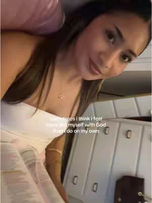 A young woman with long, dark hair is the focus. She is wearing a white top and a necklace. The background suggests an interior space, possibly a bedroom or closet, with cabinets and a pink pillow visible.