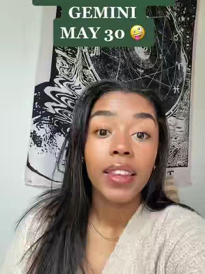 A woman with long, dark hair looks directly at the camera. Over her head is a green rectangular text overlay with the words GEMINI and MAY 30, along with a smiling face emoticon and a tongue out emoji. In the background, a black and white patterned tapestry hangs on the wall.
