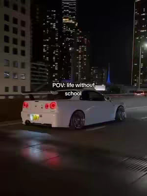 A white sports car is seen from the rear, driving on a road at night. Tall buildings with illuminated windows are visible in the background. The image has a dark, moody atmosphere, with reflections on the wet road surface.