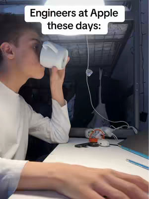 A young man in a white shirt is drinking from a white mug, with text overlayed at the top. The background is dark, featuring a desk with various items like a notepad, pen, and earphones, along with a ceiling-mounted light fixture.