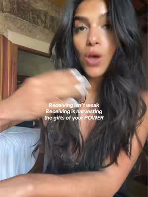 A woman with long dark hair is the central figure, her face in focus, lips slightly parted as if speaking. The background includes glimpses of a room with a stone wall and what appears to be a window or frame, enhanced by natural lighting. Text overlays appear on the image.