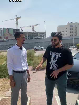 Two men are standing on a brick and concrete surface outside on a sunny day. One man is wearing a white shirt and grey pants, while the other is wearing a black Tupac T-shirt. The man in the T-shirt is wearing sunglasses and has his hand up by his stomach, gesturing with his right arm. Buildings and construction cranes are visible in the background, as well as a car parked on the side of the road.