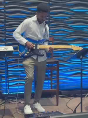A man in a white shirt and khaki pants plays a blue electric guitar in front of a blue textured wall. A music stand and a stool are nearby.