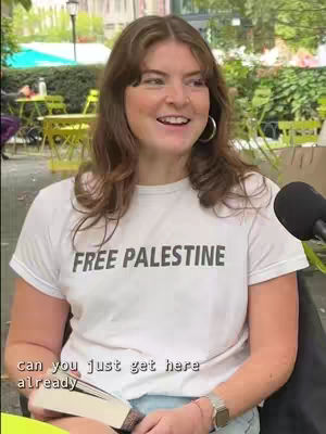A woman with long brown hair is smiling and looking at the camera. She wears a white t-shirt with the words 'FREE PALESTINE' in black letters. The background suggests an outdoor setting with green foliage and tables.
