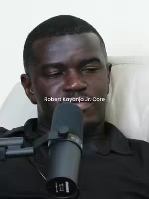 A medium shot of a man with short, dark hair and a dark complexion. The man is centered, looking slightly down with a neutral expression. A black microphone is positioned in front of his face. The background appears to be a light, neutral color. Text overlayed on the image reads: 'Robert Kayanja Jr. Core.'