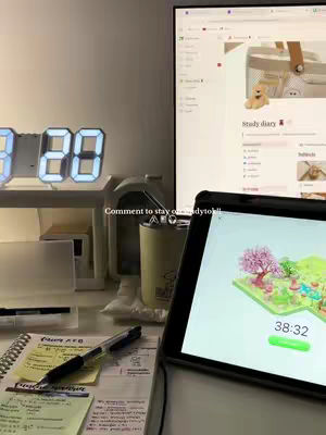 A well-lit workspace with a digital clock displaying the time, a computer monitor showing a webpage, and a tablet displaying a productivity app are visible. The scene is organized with a pen and notepad, along with a pen and a minimalist aesthetic, suggesting a focus on productivity and study.