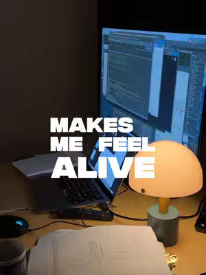 An interior shot of a desk with a laptop, a monitor displaying code, and a desk lamp. Text overlay reads: "MAKES ME FEEL ALIVE."