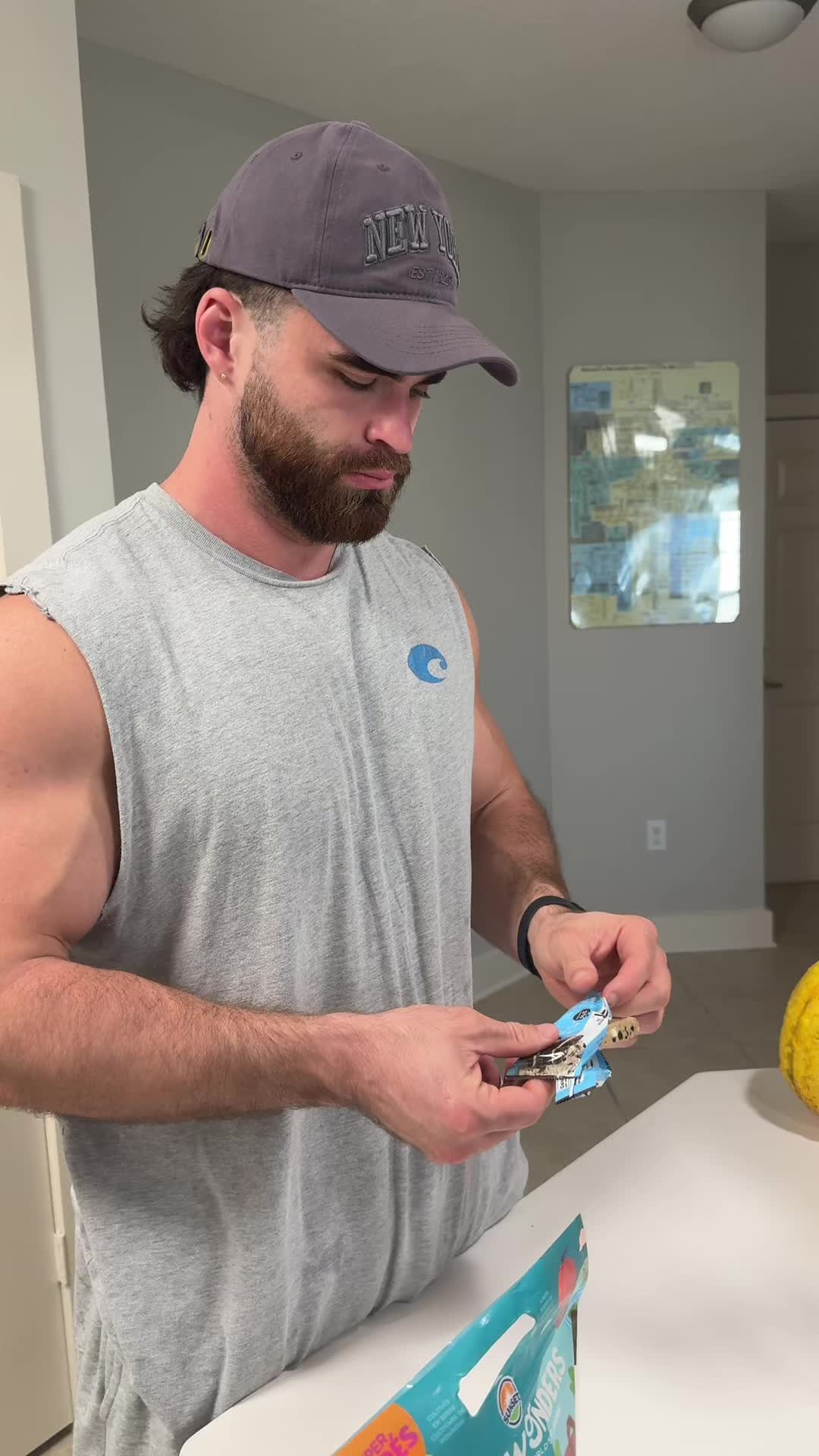A man in a gray sleeveless shirt and a New York Yankees cap stands in what appears to be a kitchen setting next to a white counter. He is looking down at a candy or snack package, holding it with both hands. The background features a light wall and a printed image or map.