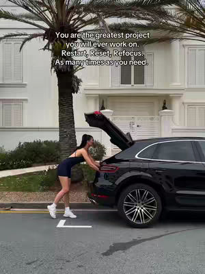 A woman dressed in athletic wear opens the trunk of a black SUV parked on a paved surface. A palm tree and a building with white walls and several windows serve as the backdrop. Text overlayed on the picture reads: 'You are the greatest project you will ever work on. Restart, Reset, Refocus. As many times as you need.'