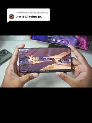 The image shows a person holding a smartphone displaying a game interface. The game appears to be a first-person shooter, with a desert-like environment visible on the screen. Hands are in the foreground, holding the phone. Visible in the background is a second phone and the surrounding environment. A comment bubble at the top contains the text 'bro is playing pc'.