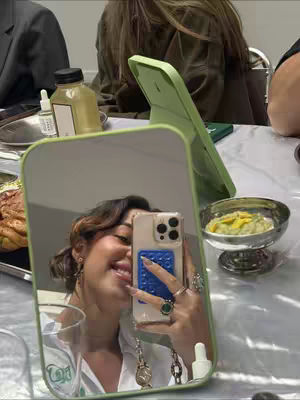 The cover image shows a mirror reflecting a woman smiling, holding her phone up to the camera. The layout features the mirror as the central element, with a table setting in the background including food and drink items. The color palette appears to be natural tones with pops of green.