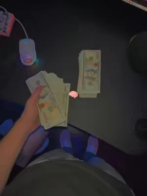 The image shows a person's lap and hands holding stacks of US dollar bills. A computer mouse and keyboard are visible in the background. The lighting is dim.