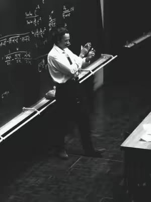 A black and white photo of a man, presumably a scientist or professor, standing and gesturing in a lecture hall setting. He is wearing a white dress shirt and dark pants, presenting in front of a chalkboard covered in complex mathematical equations. He is holding a device, possibly a slide projector. The lighting is dramatic with strong shadows, highlighting the subject and the equations on the board.