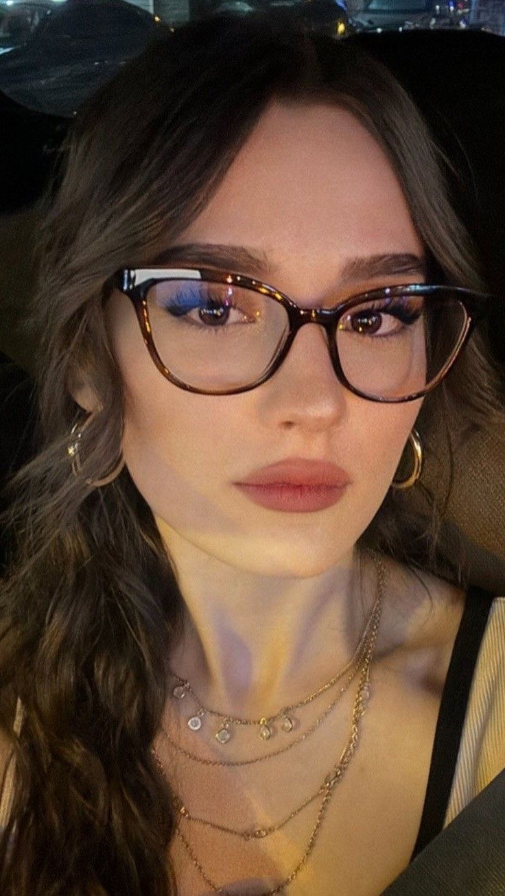 A close-up shot of a woman wearing glasses, with her face centered. She has brown hair and is wearing a few layered necklaces. She has a slight smile and a neutral expression. The background is dark, suggesting she might be in a car.