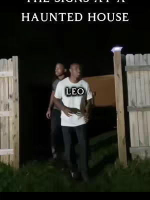 Two men standing in front of a haunted house entrance. The man in the foreground is wearing a white shirt with the word "LEO" printed on it. Behind him is another man wearing a dark color shirt. The setting appears to be at night, with a wooden fence and building in the background.