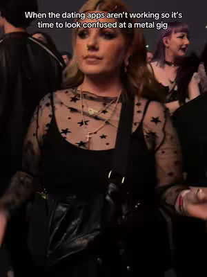 A woman stands in a crowded, dimly lit space, likely a concert. She is the primary focus, wearing dark clothing, a sheer top with star patterns, and multiple necklaces. The background blurs into the crowd, hinting at a music venue atmosphere.