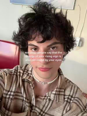 A selfie featuring a young man with dark, tousled hair and a patterned shirt. He is wearing a pearl necklace and has a message overlayed on the image.