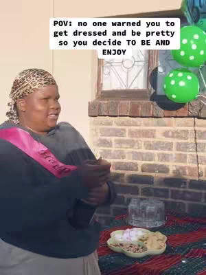 A person in a headscarf and dark top smiles while standing outside near brickwork and balloons. Text overlay at the top. A table with refreshments is in the foreground. Green polka dot balloons on the right.