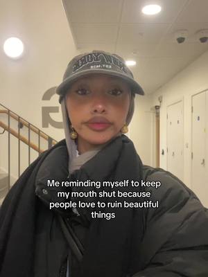 A selfie-style shot featuring a young woman. She is wearing a casual outfit with a dark, puffy jacket, and a baseball cap. The woman has a neutral expression and is looking directly at the camera. The background appears to be an indoor hallway or lobby, with a staircase, some doors, and overhead lighting. Text is overlaid at the bottom with the message: 'Me reminding myself to keep my mouth shut because people love to ruin beautiful things.'