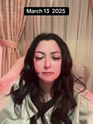 A woman with dark hair, long eyelashes and a hint of red light on one cheek is looking down with a neutral expression. Text overlay in the top center states 'March 13 2025'. The background is a light brown curtain.