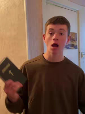 A young man stands indoors, partially visible. He faces the camera with his mouth open, as if speaking. He holds a book with a cross on it in his left hand.. The background includes parts of a door, a light-colored wall, and glimpses of interior design. The man is wearing a dark long-sleeved shirt.