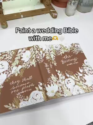 The image shows an open, decorated Bible with floral designs and calligraphy on a light surface, possibly a table. In the background, there's a wooden easel and some blurred items, suggesting an art or craft setting.